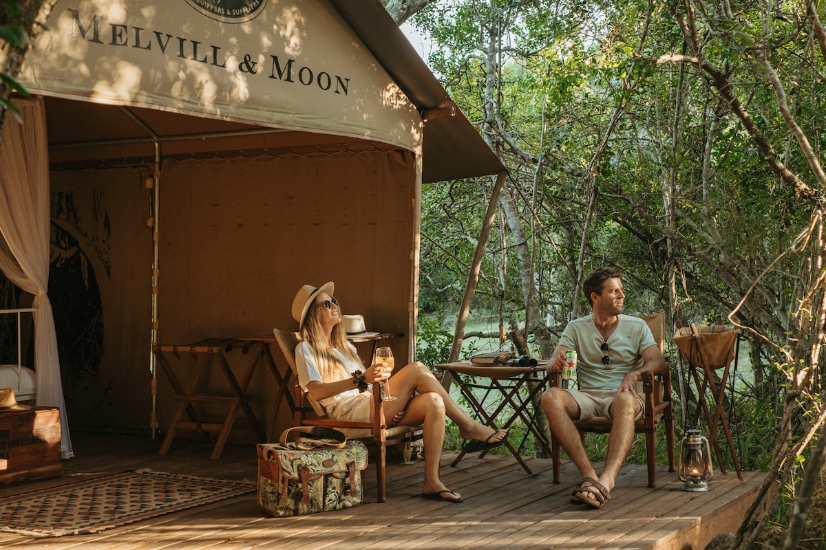 A canvas tent features a wooden deck surrounded by lush greenery. Two individuals are seated on comfortable chairs, enjoying beverages. A rustic table and lantern are positioned nearby, enhancing the outdoor experience amidst the natural surroundings.