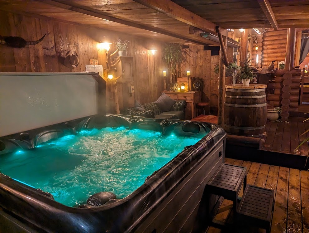 A hot tub is prominently featured, surrounded by wooden walls and ambient lighting. The water glows in a calming turquoise hue, while potted plants and decorative elements add to the rustic charm of the space. Nearby seating is provided for relaxation.