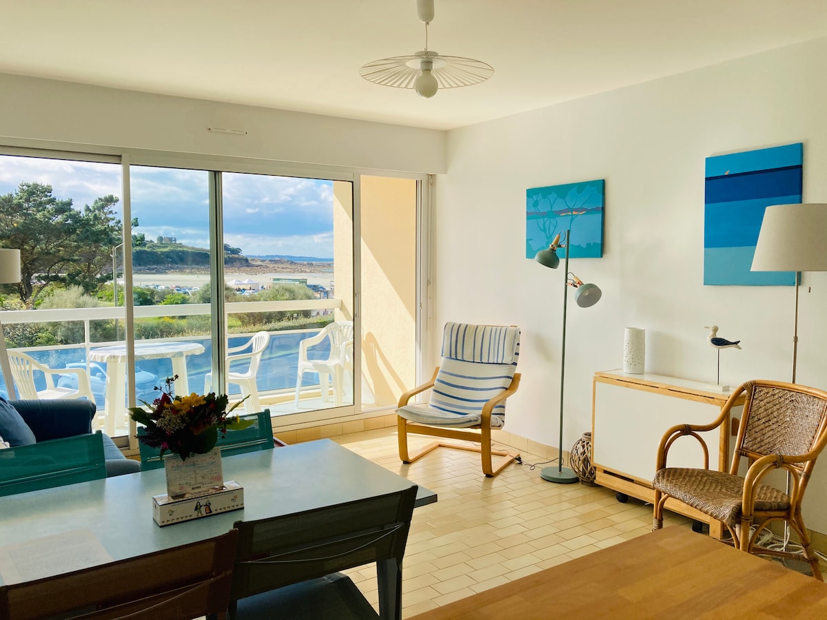 A bright living area features large sliding glass doors that open to a balcony with a view of the beach. A wooden dining table sits beside a cozy armchair, while wall art in calming blue tones complements the light, airy space.