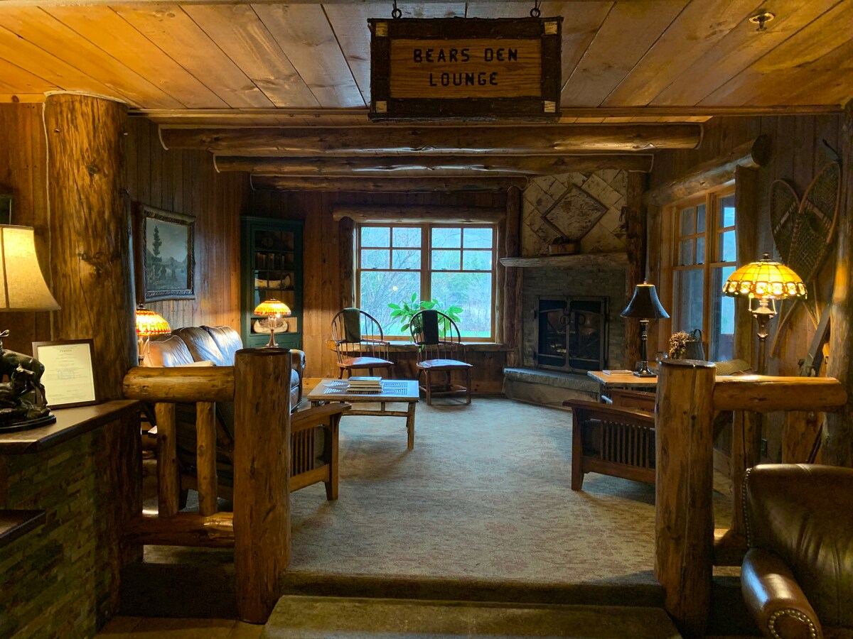 A cozy lounge area features warm wooden walls and rustic furnishings. Two rocking chairs sit near a large window, allowing natural light to fill the space. A gas fireplace is flanked by two lamps, and a wooden sign reading 'Bears Den Lounge' hangs above.