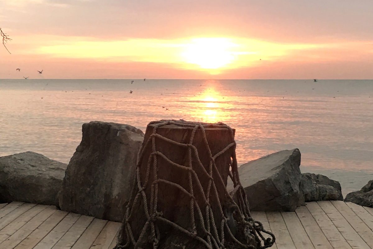 A serene view of the sunset reflecting on calm waters is featured, with soft orange and pink hues filling the sky. A rustic, rope-bound structure sits prominently in the foreground, framed by smooth stones, enhancing the natural lakeside setting.