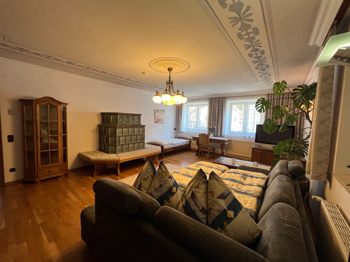 A spacious living room features a large, comfortable sectional sofa with decorative cushions. Sunlight fills the room through expansive windows, highlighting a dining table and a sideboard. A cozy seating area is complemented by a large plant, contributing to a relaxing environment.