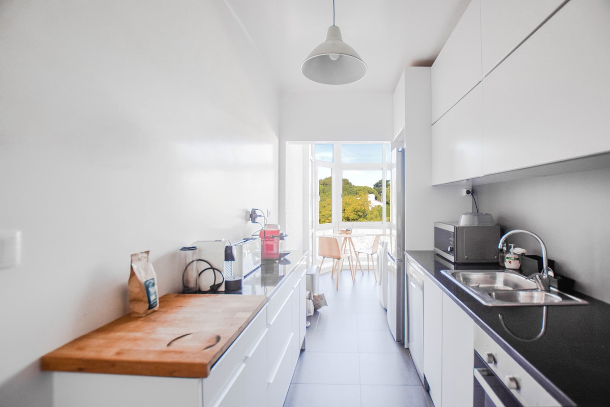 A modern kitchen is shown, featuring white cabinetry and a wooden countertop. A sink and cooking area provide functionality, while large windows offer natural light and views of greenery outside. A small dining table with chairs is visible in the distance.