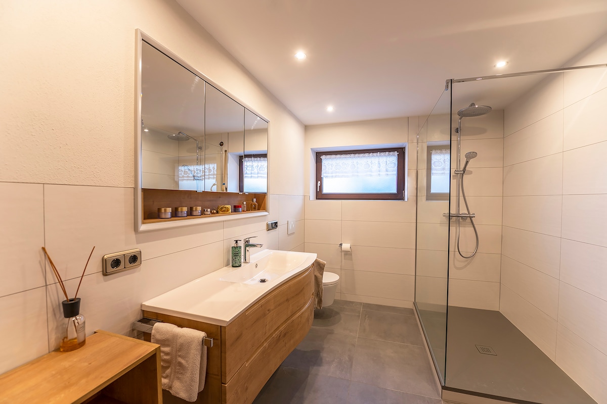 A modern bathroom is presented with a spacious shower area enclosed in glass. A wooden vanity with a large mirror hangs above, providing ample storage. Subtle lighting enhances the neutral tile accents, while a towel is neatly placed on the vanity. Natural light filters through the nearby window.