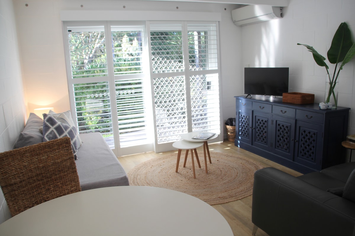 The living room features a light, airy design with a single day bed and a two-seater leather lounge. A small round table sits on a natural fibre rug, complemented by a dark blue cabinet and a flat-screen Smart TV. Outdoor greenery is visible through the large windows.