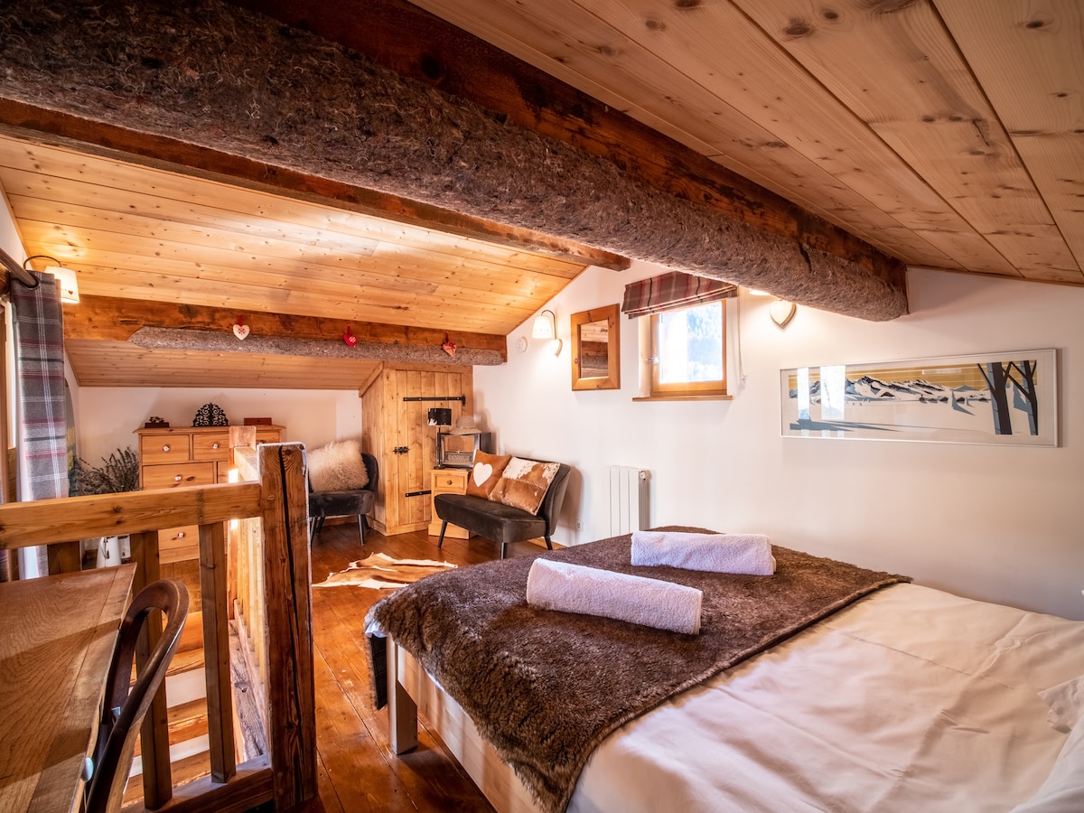 An upper-level sleeping area features natural wood beams and a cozy, warm feel. A spacious bed is adorned with soft linens and two rolled towels. Accents include a wooden dresser, a comfortable chair, and a window offering light, adding to the inviting ambiance.