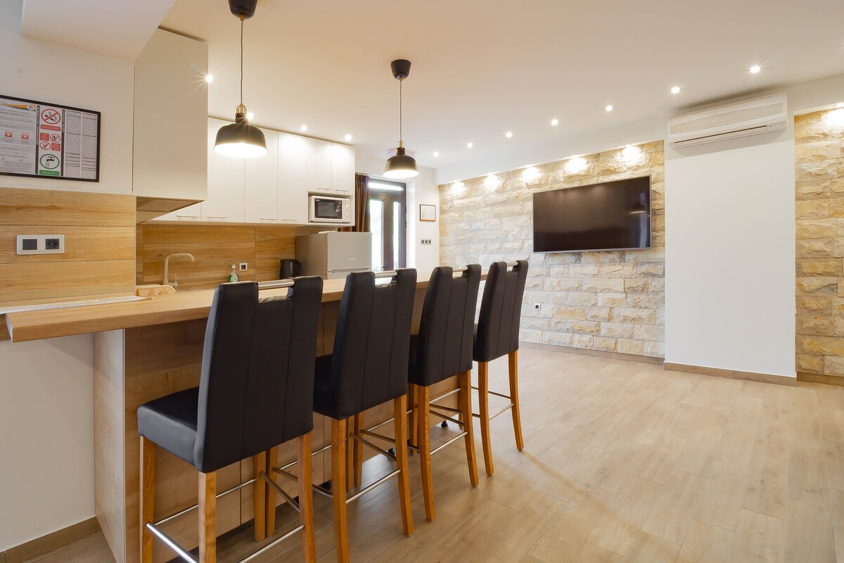 A modern kitchenette features a stylish bar with high stools, providing ample seating. Under-cabinet lighting highlights the wood accents, while stone walls create a warm contrast. A flat-screen TV is mounted on the wall opposite, and natural light fills the open living area.