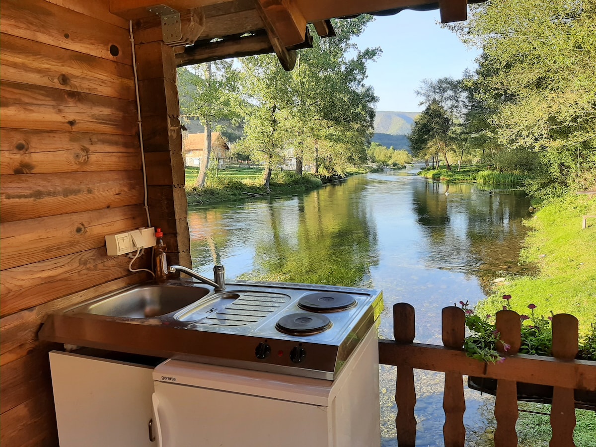 An outdoor kitchen area is featured with a stainless steel sink and a two-burner stove, set against a backdrop of a serene river. Lush greenery surrounds the water, and a wooden fence frames the scene, providing a natural and tranquil setting.