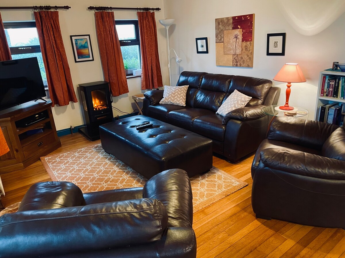 A cozy sitting room is featured, furnished with dark leather couches and a large ottoman. A stylish electric stove adds warmth, while a smart TV sits on a wooden console. Light filters through windows adorned with curtains, and wall art enhances the inviting space.