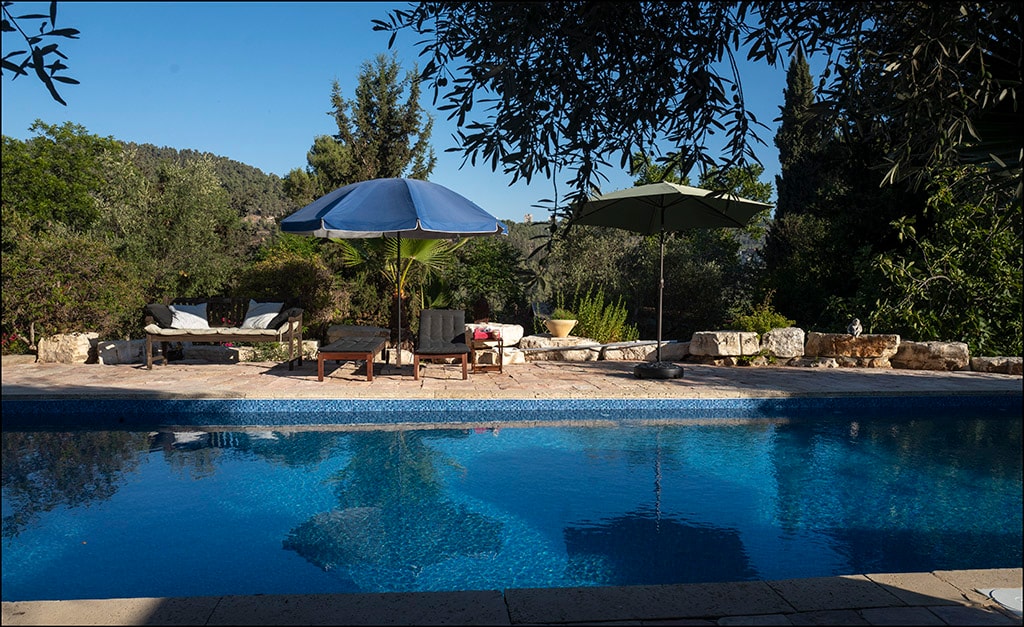 The private saltwater pool reflects clear blue skies and is surrounded by lush greenery, creating a serene outdoor space. Chaise lounges and umbrellas are positioned on the pool terrace, providing shaded seating areas for relaxation.