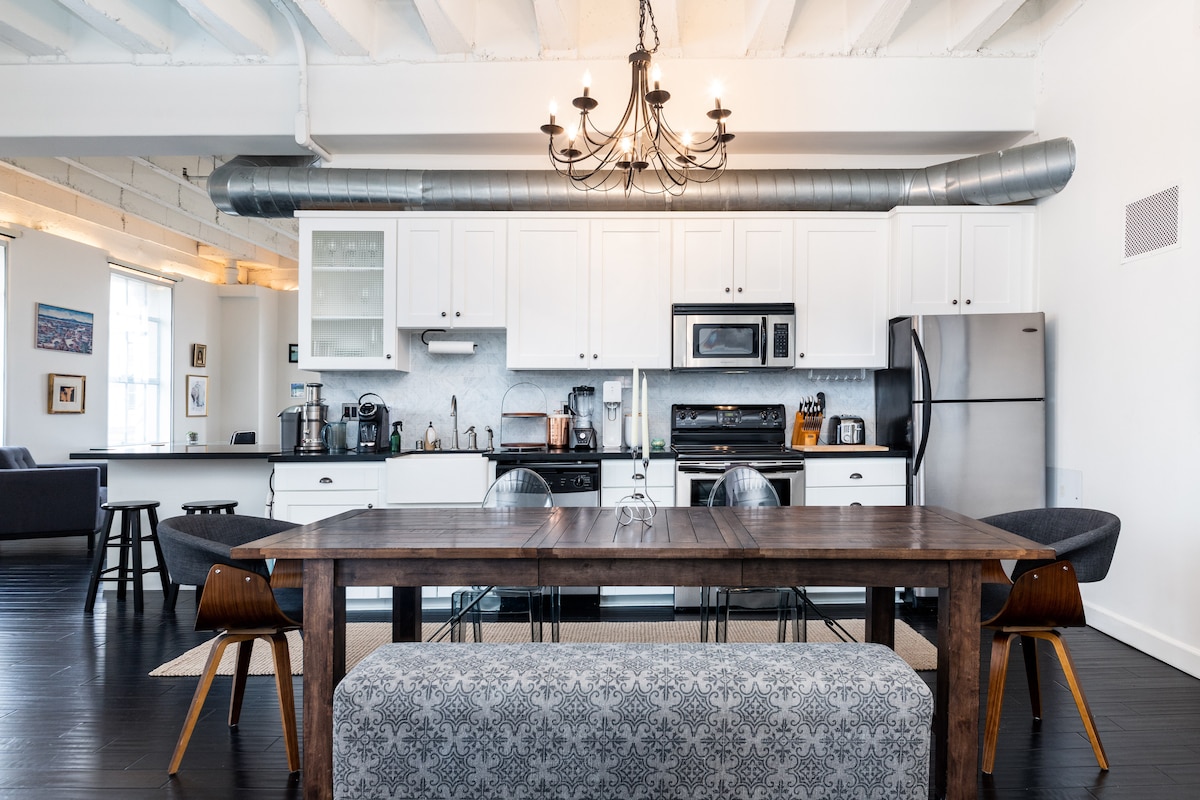 The open kitchen and dining area features a large wooden table surrounded by modern chairs. A well-equipped kitchen is visible in the background, complete with a refrigerator, microwave, and various appliances. Soft natural light illuminates the space through multiple windows.