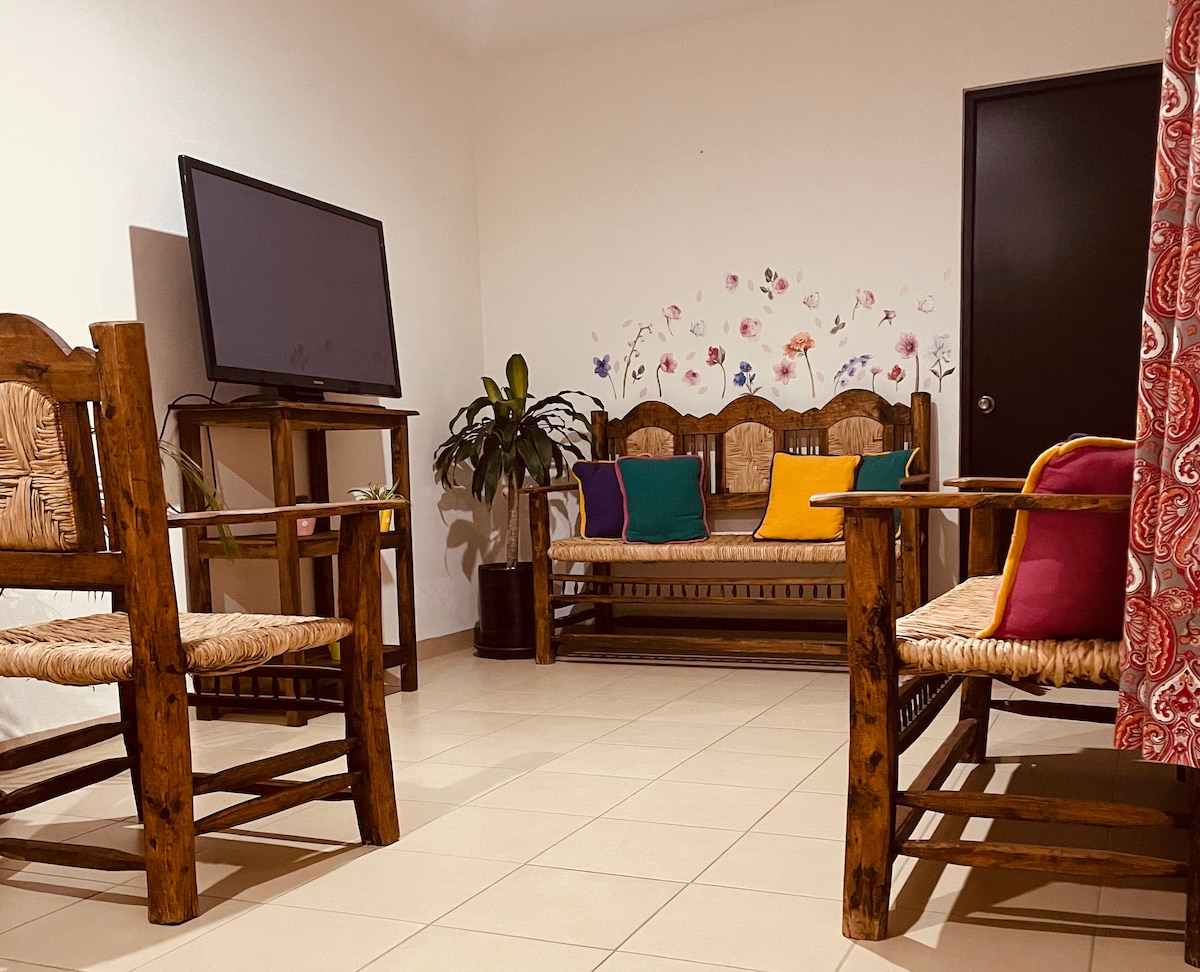 A welcoming living area is furnished with rustic wooden seats adorned with colorful cushions. A flat-screen television is positioned on a stand, with a decorative plant adding a touch of greenery. The walls feature subtle floral artwork, enhancing the space's charm.