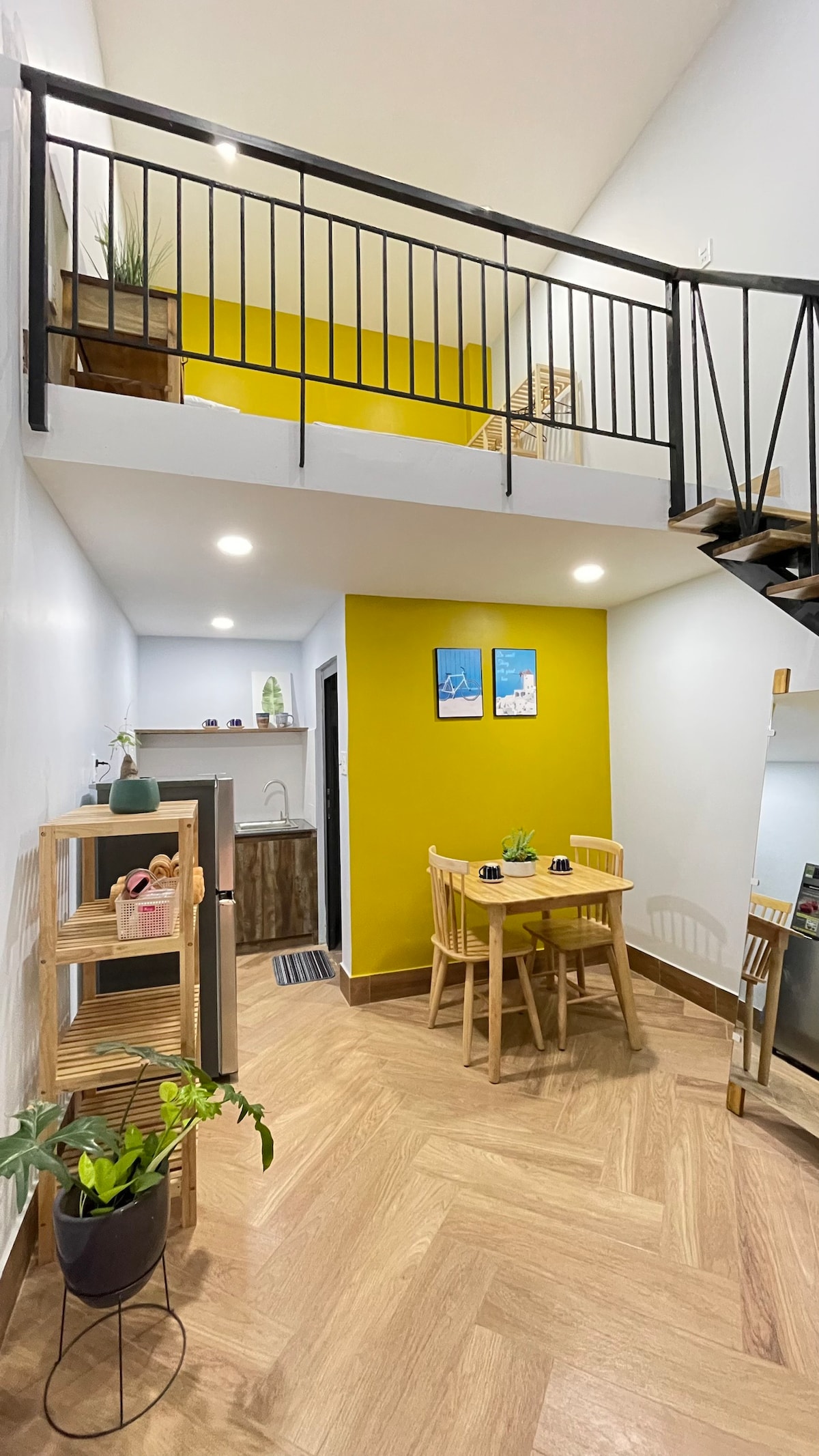 A bright studio space features warm yellow walls and wooden accents. A compact dining area is set with a small table and two chairs. A staircase leads to an upper loft area, and plants are arranged on shelves, adding a touch of greenery.