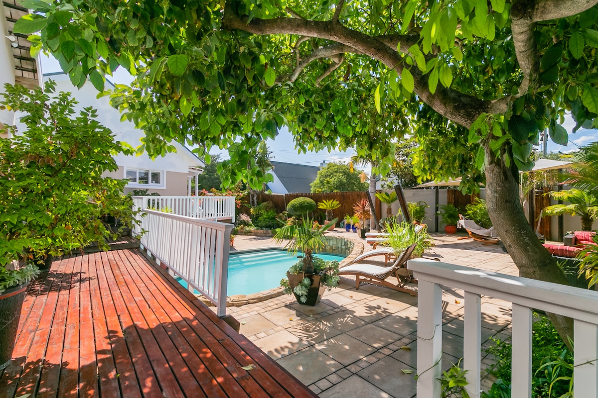 A sunlit outdoor area features a refreshing swimming pool surrounded by lush greenery. A wooden deck is visible alongside the pool, with lounge chairs positioned for relaxation. The scene is framed by a large tree, providing shade and enhancing the tranquil setting.