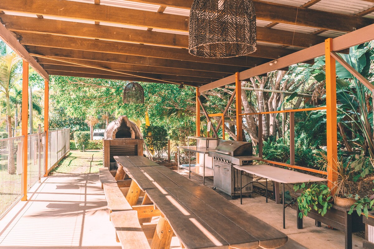 A spacious outdoor covered area features long picnic-style tables and a gas barbecue station. Abundant greenery surrounds the space, providing a natural ambiance. Two woven light fixtures hang from the ceiling, offering a rustic touch to the inviting gathering area.