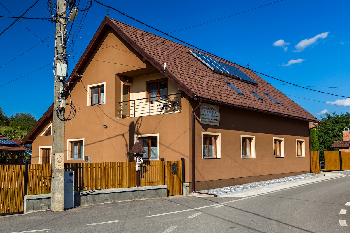 The exterior of a two-story building features a warm brown façade complemented by a sloped roof. Solar panels are installed on the roof. The property is bordered by a wooden fence, and the surrounding area is clear with a road visible on one side.