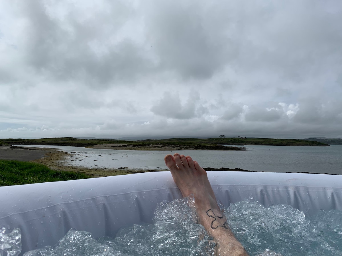 A foot is seen resting on the edge of a bubbling hot tub, with water cascading around it. The view encompasses a cloudy sky and a serene landscape of grassy hills and a distant shoreline along the water's edge.