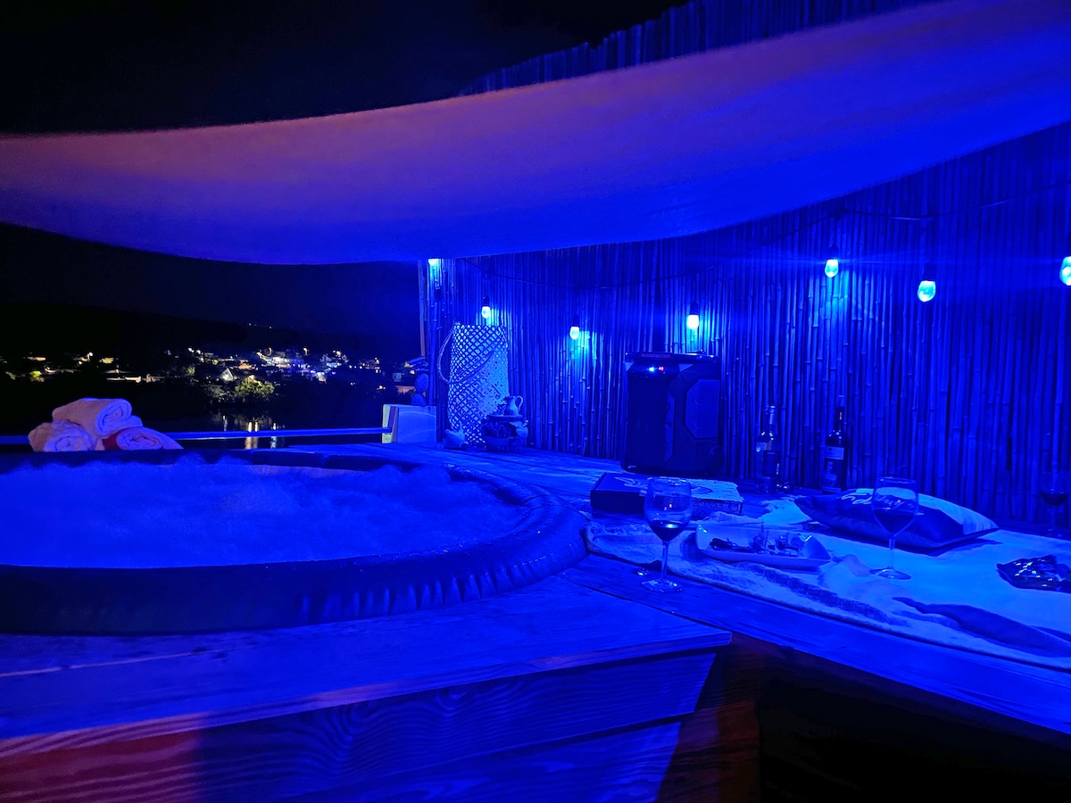 A private terrace is illuminated by soft blue lights, featuring a jacuzzi surrounded by wooden decking. A table is set with refreshments, and bottles of wine are present. The distant view reveals twinkling lights from the surrounding area, adding to the serene evening atmosphere.