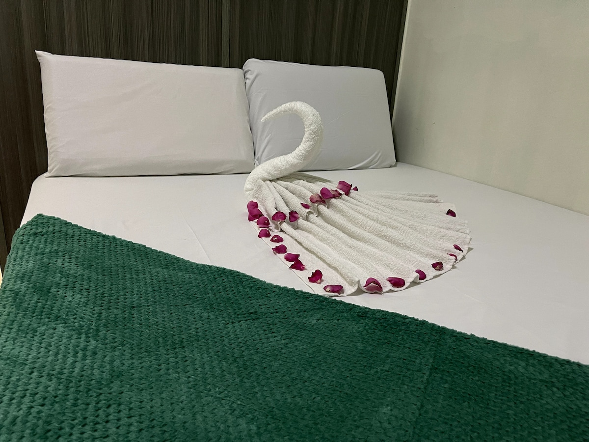 A neatly made bed features two comfortable pillows and a decorative towel swan arranged with flower petals. A soft green throw blanket is positioned at the foot of the bed, complementing the overall clean and inviting appearance of the sleeping area.