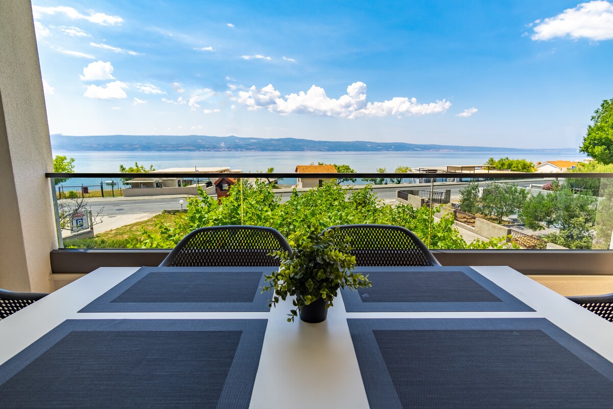A balcony is depicted with a dining table surrounded by chairs, offering a view of a scenic landscape. Lush greenery and water can be seen in the background under a wide sky, creating a serene outdoor space.