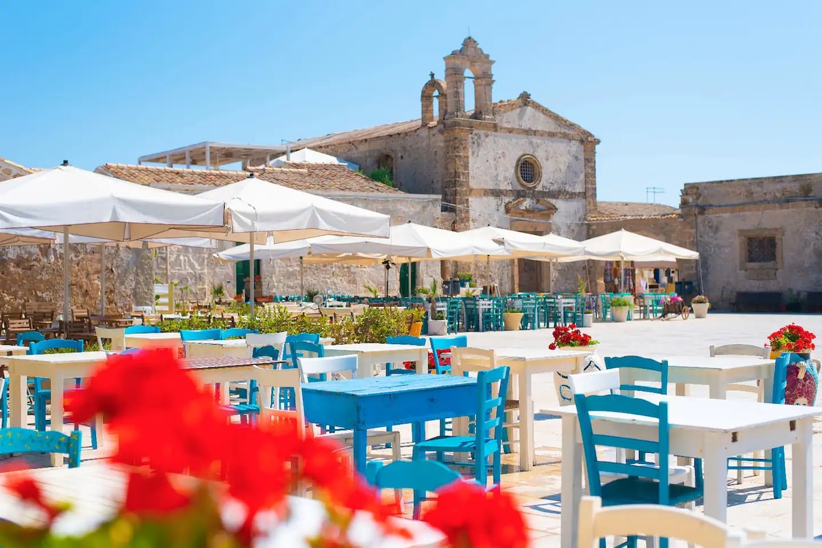 A spacious outdoor area is presented, featuring several white umbrellas shading colorful tables and chairs in hues of blue and white. Bright red flowers in pots add vibrancy to the scene, and a historic building is visible in the background.