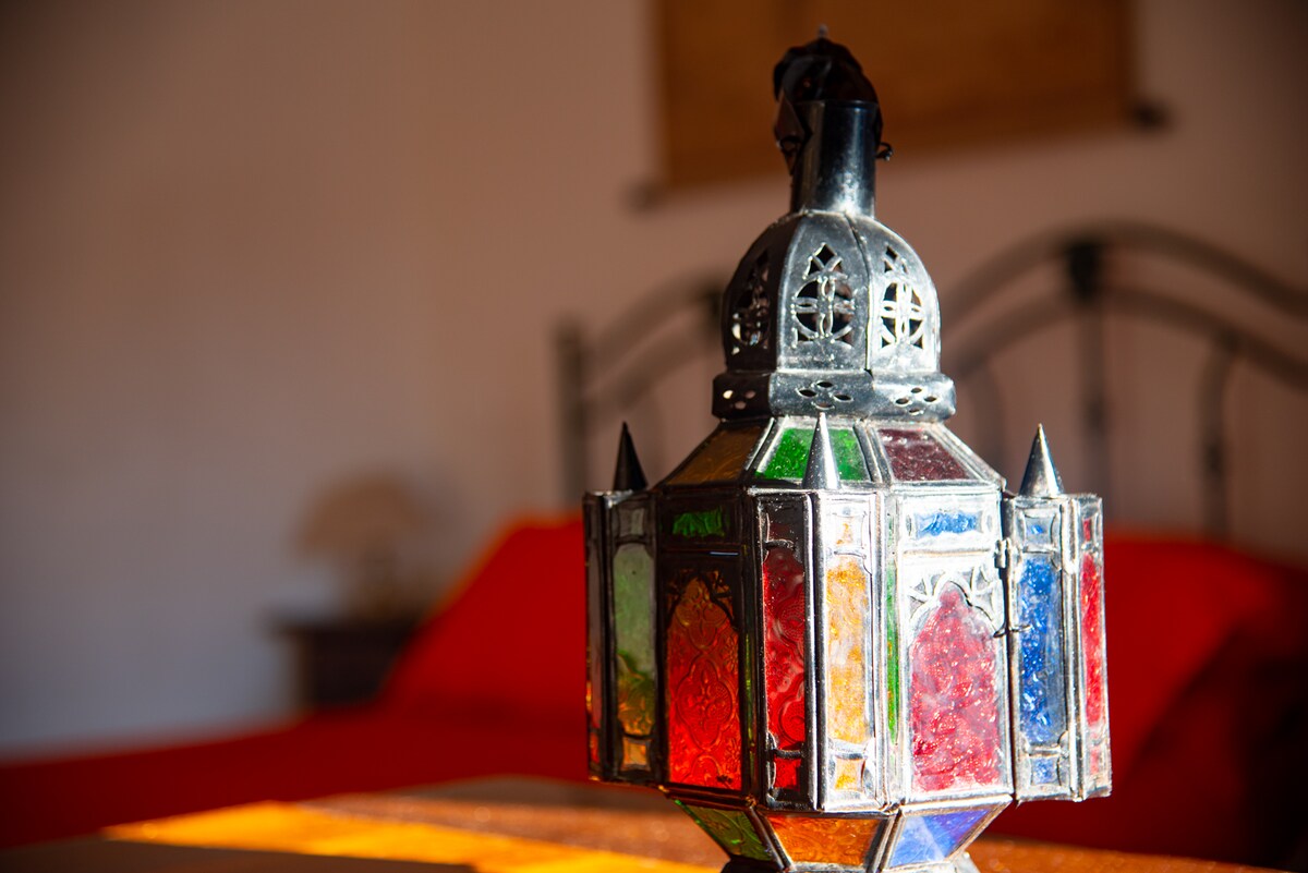A decorative lantern with colorful glass panels is centrally placed on a wooden table. The rich hues of red, green, and blue glass are highlighted by natural light, creating a vibrant focal point in the space, with a blurred background featuring a bed with red bedding.