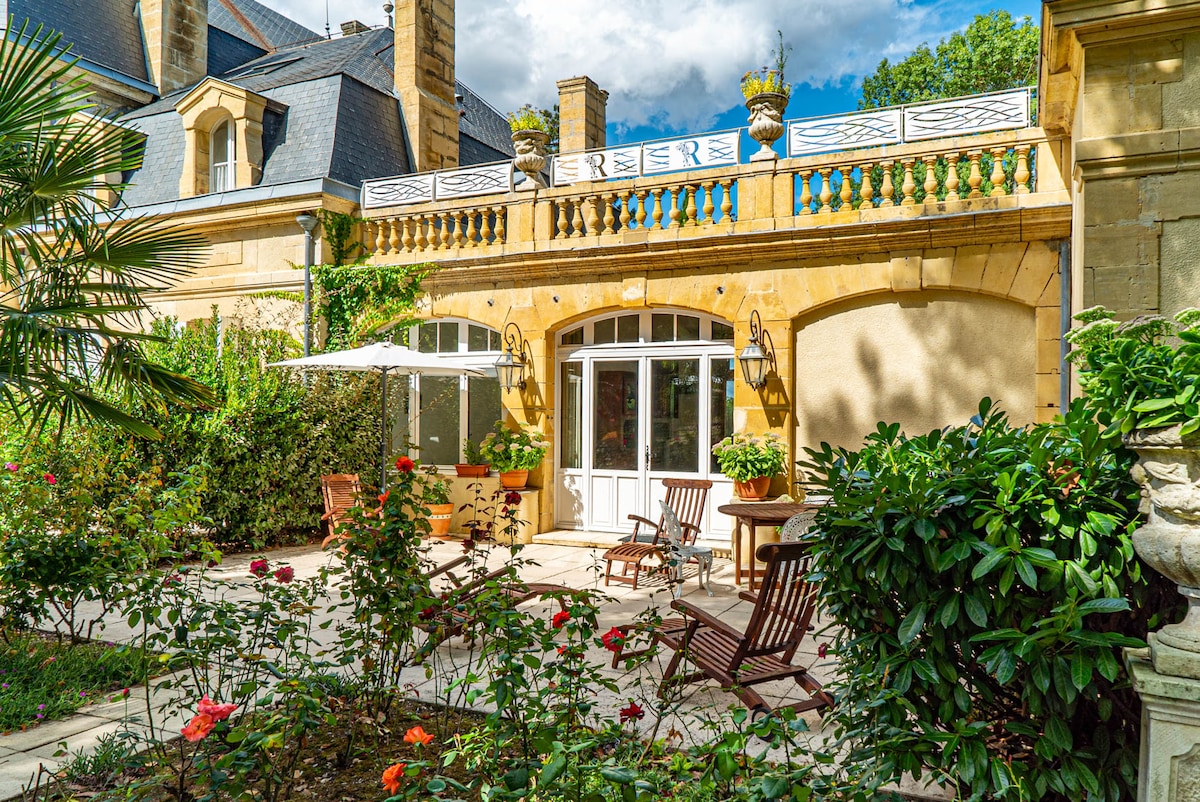 A charming outdoor terrace is framed by blooming roses and lush greenery. Wooden chairs are arranged around a small table, with an umbrella providing shade. The building's elegant facade features large glass doors that connect the terrace to the interior space.