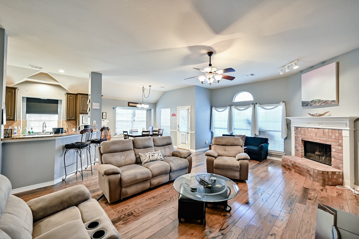 The open living area features a cozy arrangement of seating with two reclining sofas and an armchair, positioned around a coffee table. Large windows provide natural light, while a brick fireplace adds warmth. A spacious kitchen is visible in the background.