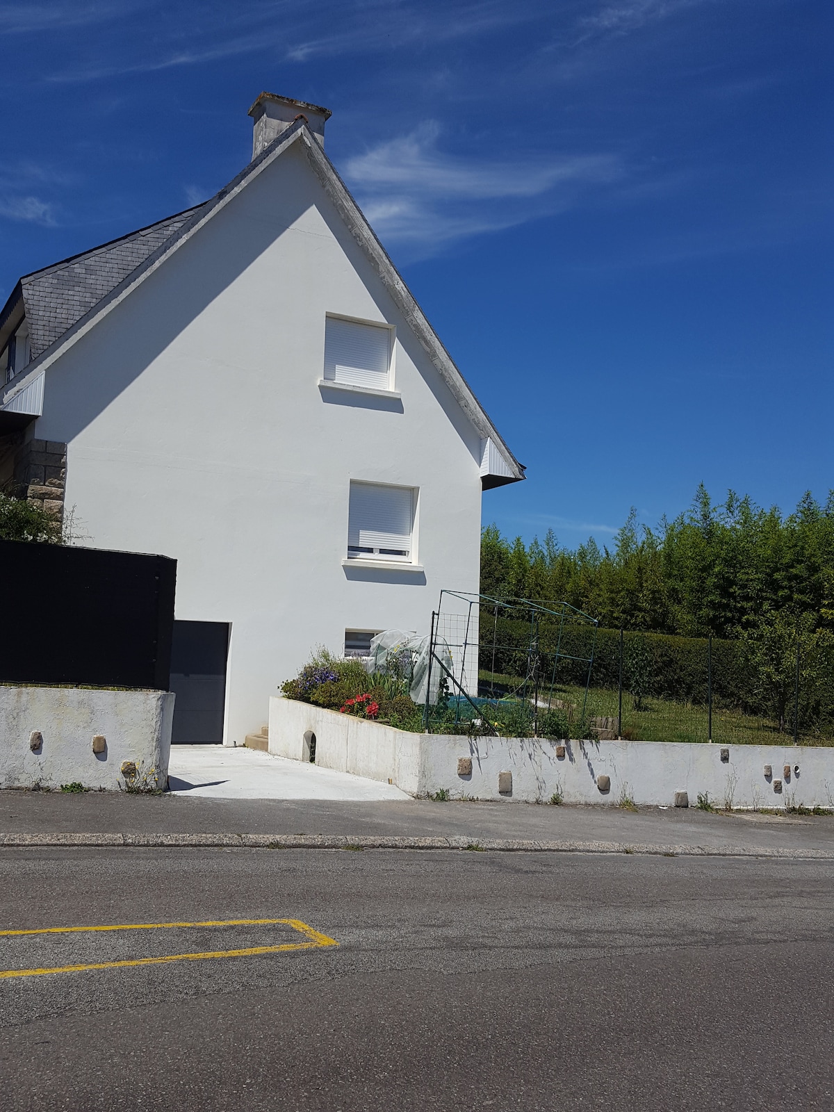 The exterior of the structure is presented, featuring a white facade with a pitched roof. A concrete pathway leads to the entrance, flanked by a small garden area with greenery. A fence surrounds the garden, and the clear blue sky above enhances the overall scene.