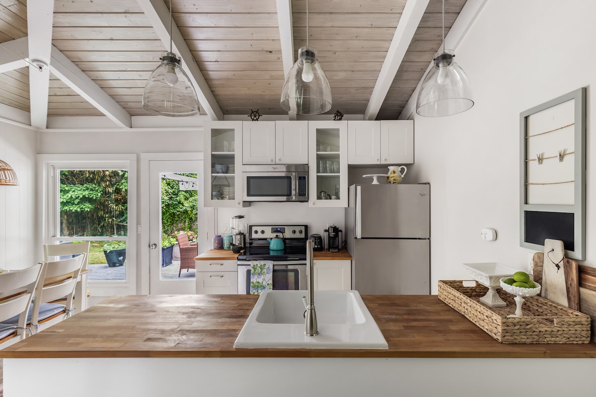 A clean eat-in kitchen features a spacious wooden island with a sink and a basket for storing items. Modern appliances, including a stainless steel refrigerator and microwave, are visible. Natural light enters through glass doors, illuminating the open space.