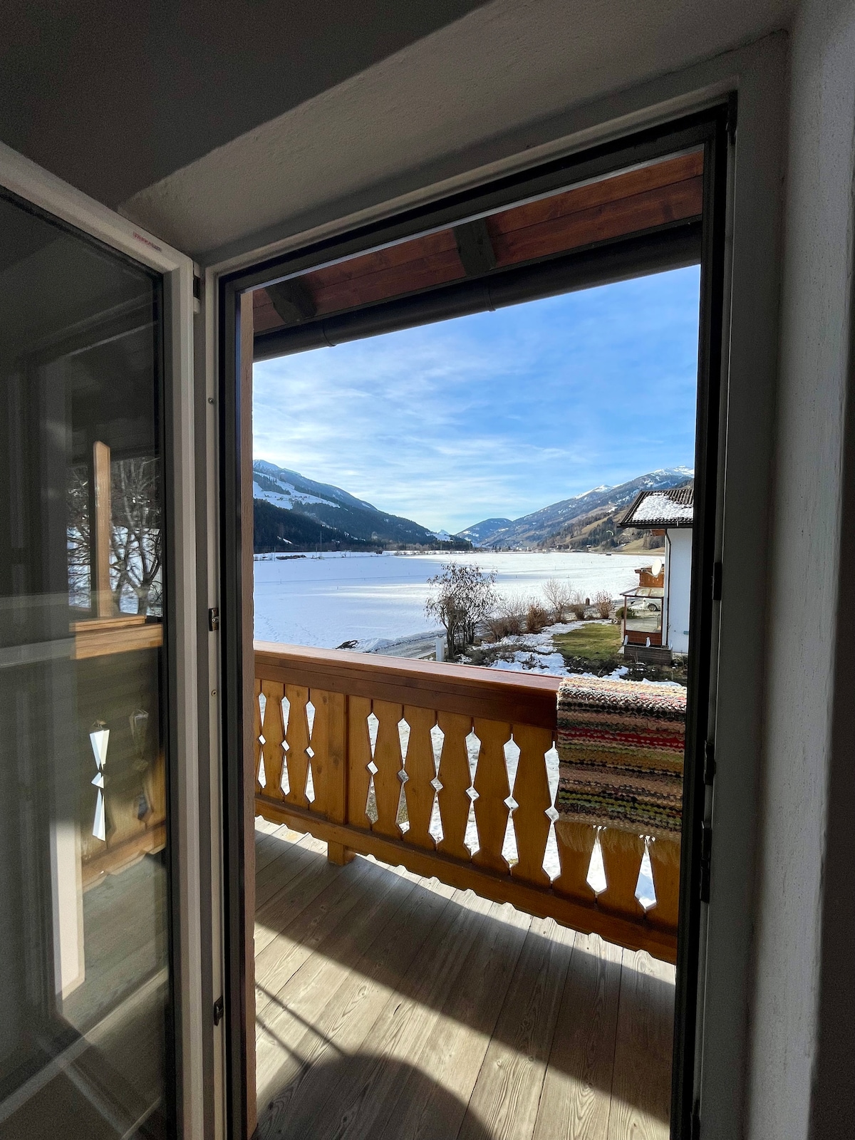 A view of a serene landscape is framed by an open door. A wooden railing borders the balcony, which overlooks a snow-covered valley and distant mountains. A woven blanket is draped over the railing, enhancing the peaceful ambiance of the setting.