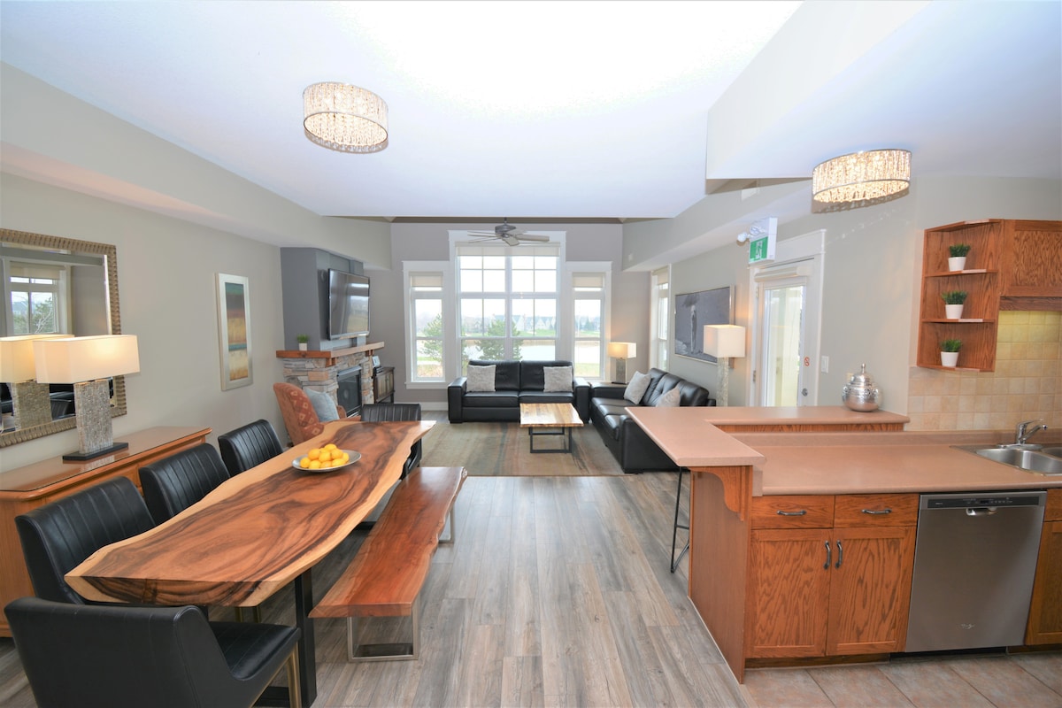 A spacious living and dining area features a natural wood dining table set for six. The modern kitchen connects seamlessly with the open layout, while a comfortable seating arrangement faces a gas fireplace. Large windows provide ample natural light and views of the surrounding landscape.