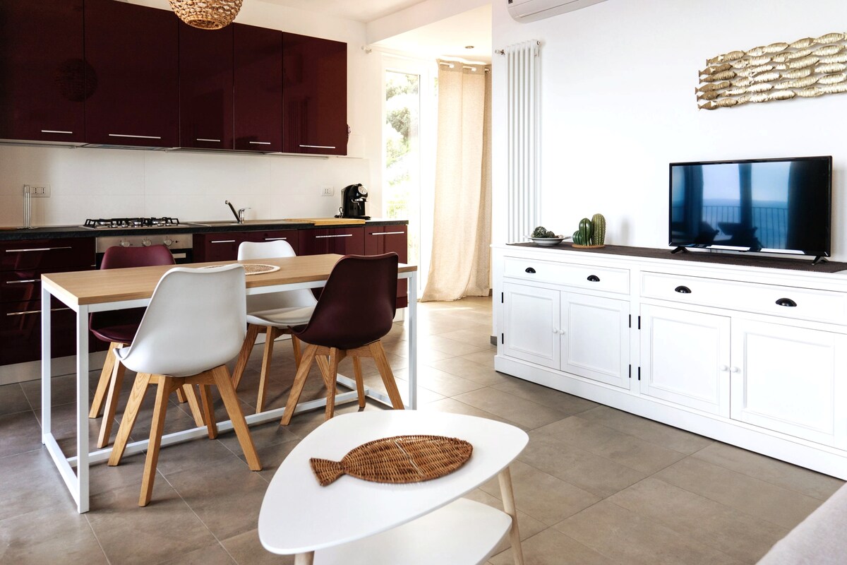 The open-plan living area is equipped with a modern kitchen featuring burgundy cabinets and stainless steel appliances. A dining table with four chairs is positioned centrally, with a white sideboard and television present nearby. Natural light enters through a large window with sheer curtains.