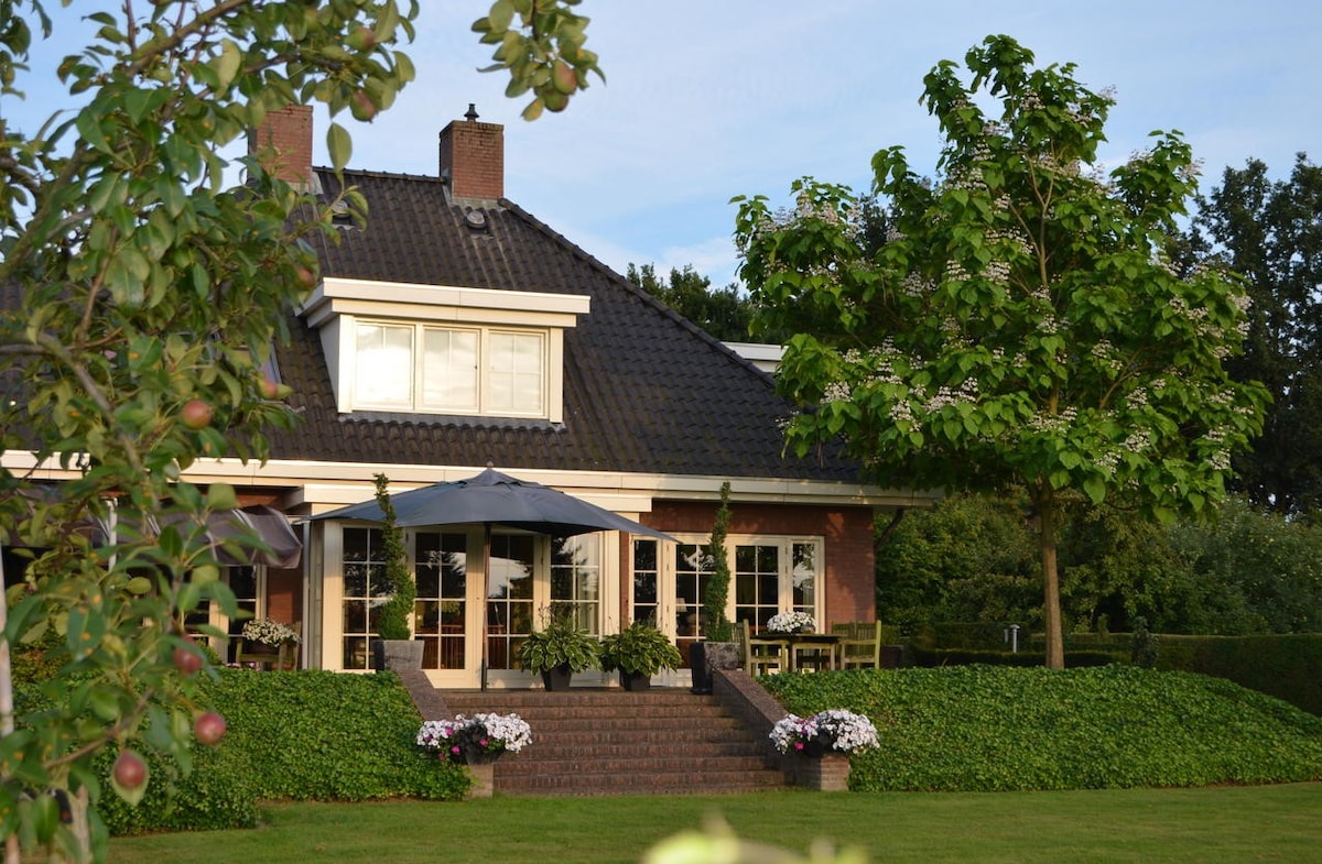 A luxurious country house is framed by lush greenery and blooming flowers. A spacious terrace with outdoor seating and umbrellas is visible at the entrance, complemented by large windows that invite natural light into the interior.
