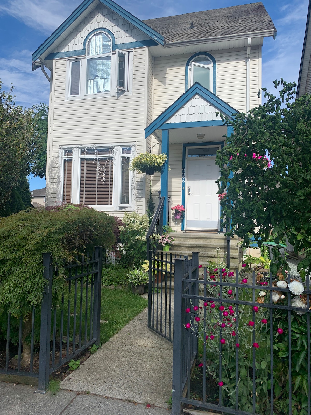 The exterior of the property is framed by a black wrought-iron fence and colorful flower beds. A set of stone steps leads to a bright front door, adorned with a small decorative wreath, while flowering plants hang from the windowsills.