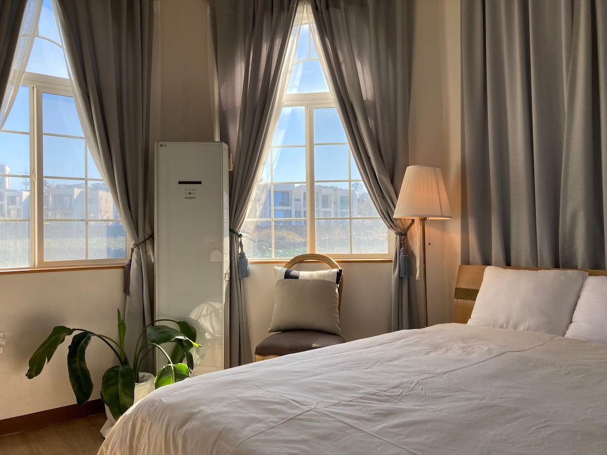 A well-lit sleeping area is shown with a spacious bed covered in crisp white linens. A chair is positioned next to a window adorned with soft curtains, allowing natural light to fill the space. A small plant adds a touch of greenery.