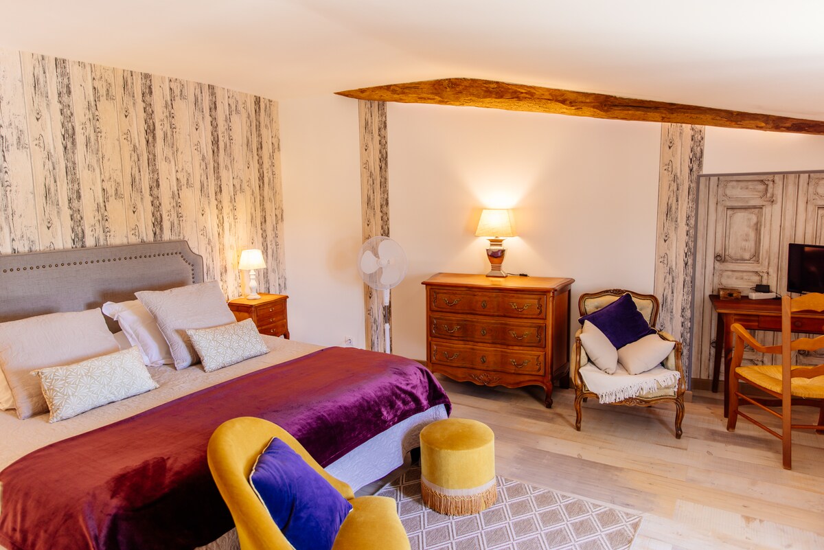 The spacious bedroom features a king-size bed with a plush purple throw, complemented by bedside lamps and wooden furniture. An antique dresser and a comfortable chair add character, while warm lighting creates a cozy atmosphere. A fan stands to the side for added convenience.