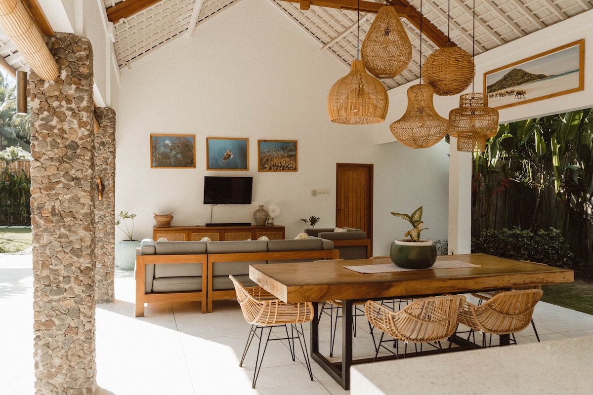 An airy living and dining area features high ceilings and natural light filtering through large windows. A wooden dining table is surrounded by woven chairs, while a comfortable seating arrangement faces a wall-mounted TV. Tropical plants and art decorate the space, enhancing the relaxed atmosphere.