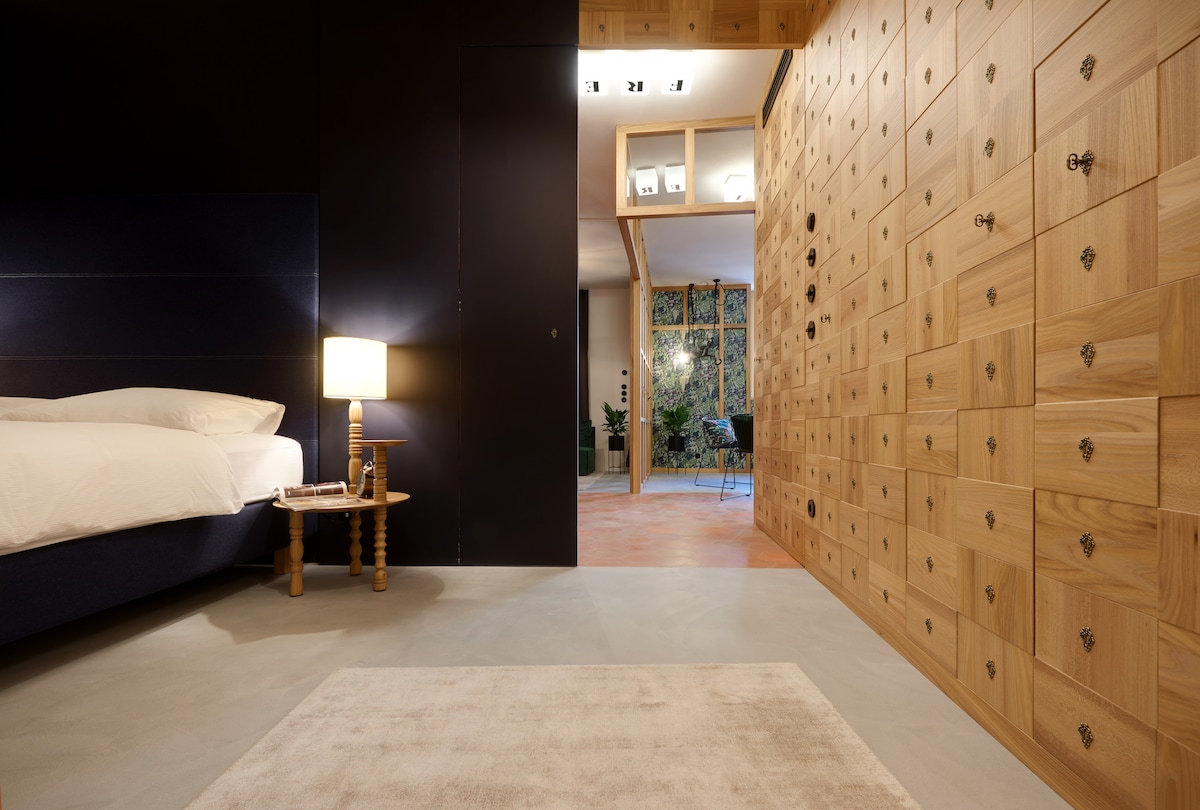 A modern bedroom is presented with a spacious bed dressed in white linens. A wooden nightstand beside the bed holds a lamp. The wall features a row of wooden drawers, contributing to a minimalist aesthetic. Natural light is visible through an opening leading to a garden.
