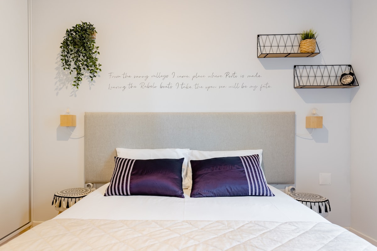 A spacious bed features a light grey headboard and is adorned with white and purple pillows. Decorative wall shelves with potted plants provide a touch of greenery. Soft lighting is offered by bedside sconces on either side, complemented by a neutral color palette.