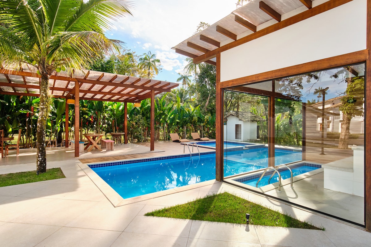 An outdoor area features a modern pool surrounded by tropical greenery. Pool loungers are arranged under a wooden pergola, while a spacious deck enhances the area. A glass wall reflects the inviting blue of the water, creating a seamless flow between the spaces.