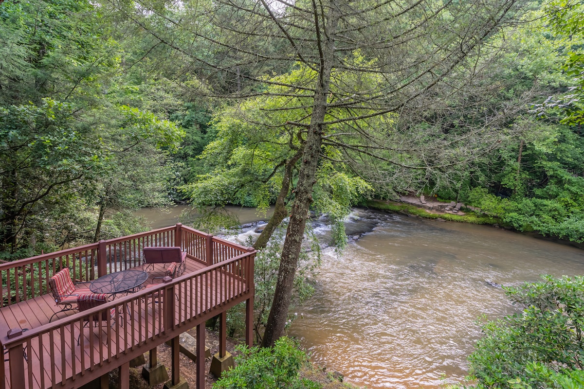 A deck overlooks a gently flowing creek, surrounded by lush greenery. A table and chairs are positioned for outdoor relaxation, inviting guests to enjoy the serene atmosphere. The sound of water enhances the tranquil setting, with trees providing natural shade.