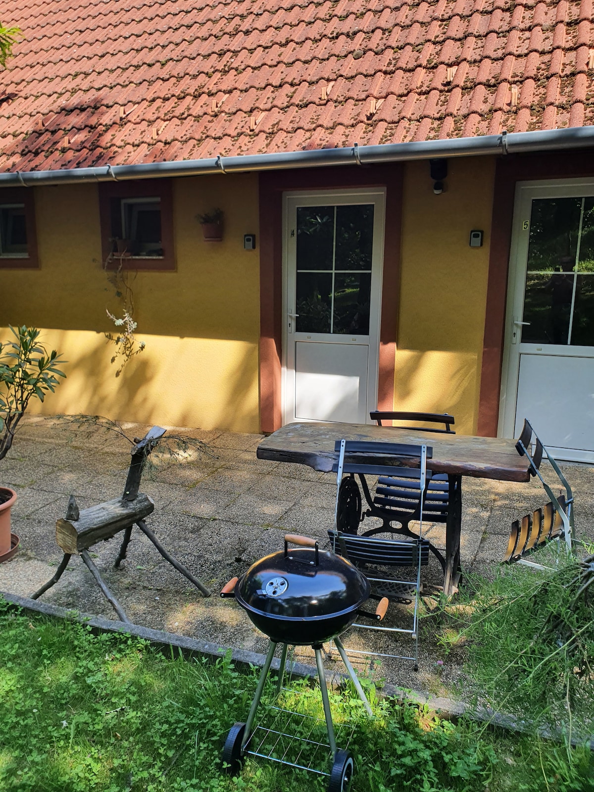 A patio area is visible, featuring a circular charcoal grill and a rustic wooden table surrounded by four chairs. The backdrop consists of a yellow wall with two doors and small windows, complemented by greenery and potted plants.
