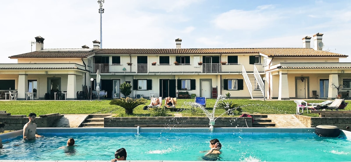 A multi-unit building showcases a spacious outdoor area with a large pool. Several guests are enjoying the water, while others relax on sun loungers nearby. Lush green grass surrounds the pool, creating a serene environment for relaxation.