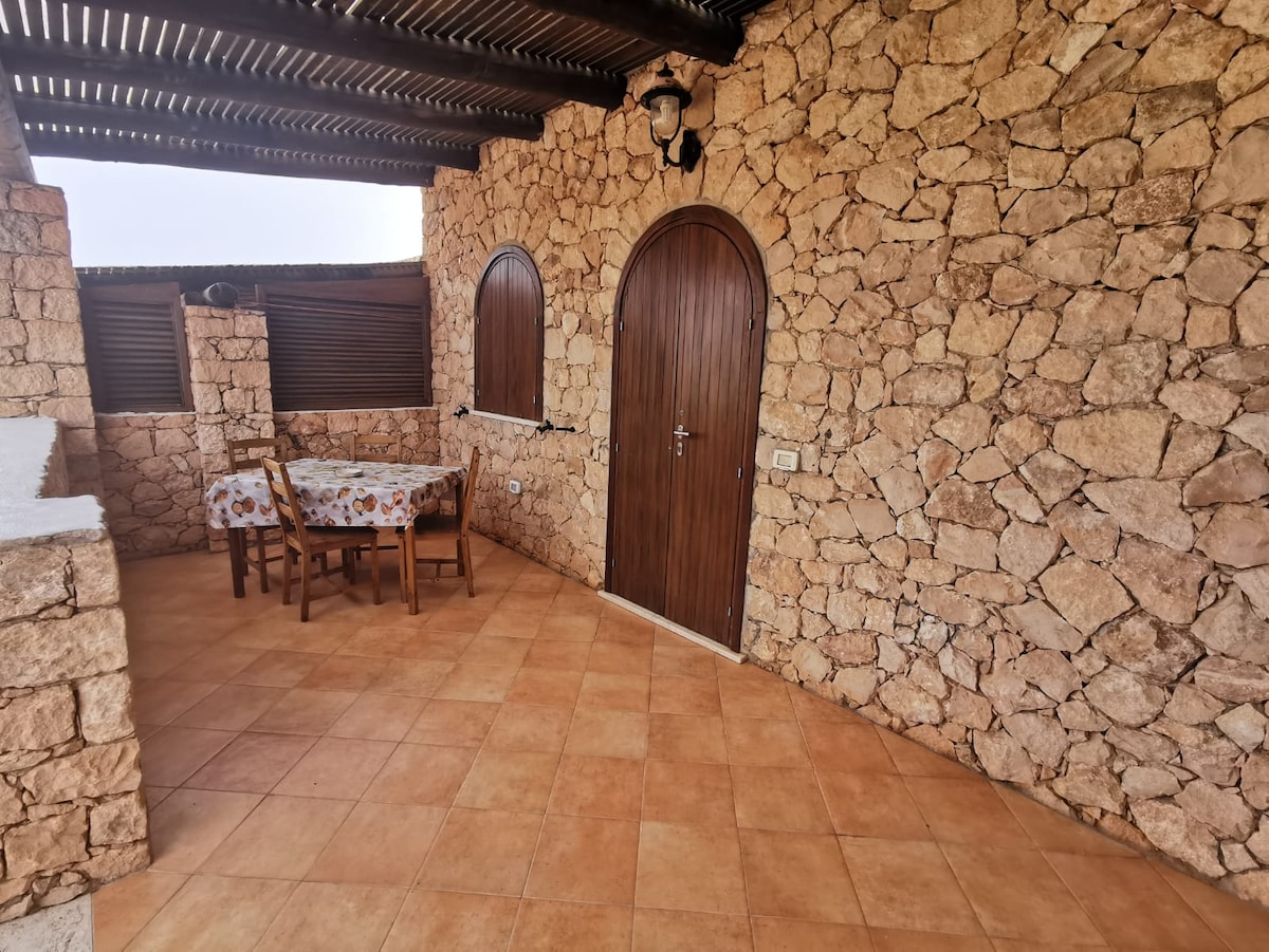 A stone-walled terrace area is featured, furnished with a wooden dining table surrounded by four chairs. A large doorway made of wood is visible, and warm, earthy tones fill the floor, enhancing the outdoor atmosphere.