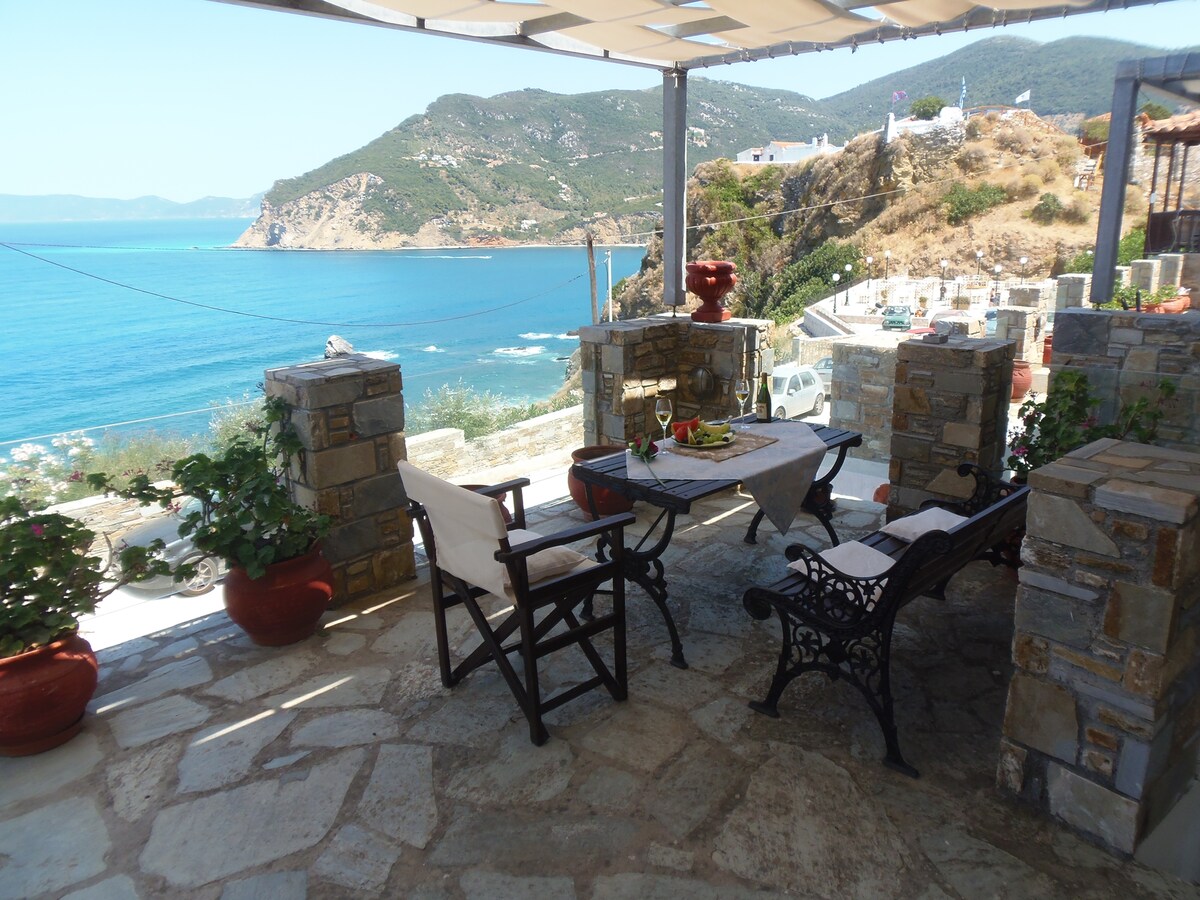 A shaded outdoor seating area is presented with a dining table and chairs, offering panoramic views of the Aegean Sea and surrounding hills. Stone and wooden features create a rustic ambiance, while potted plants add a touch of nature to the space.