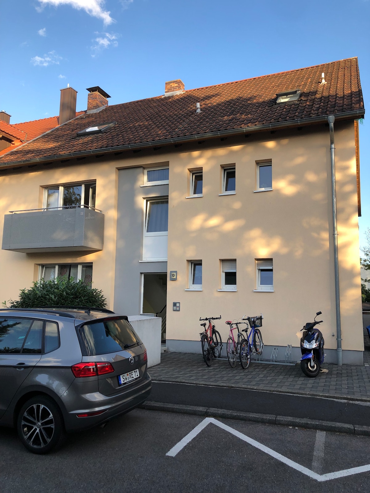 The exterior of a three-story building is shown, featuring a light-colored facade with multiple windows and a small balcony. Two bicycles are parked alongside a scooter, and a silver car is positioned in front of the building on a paved area.