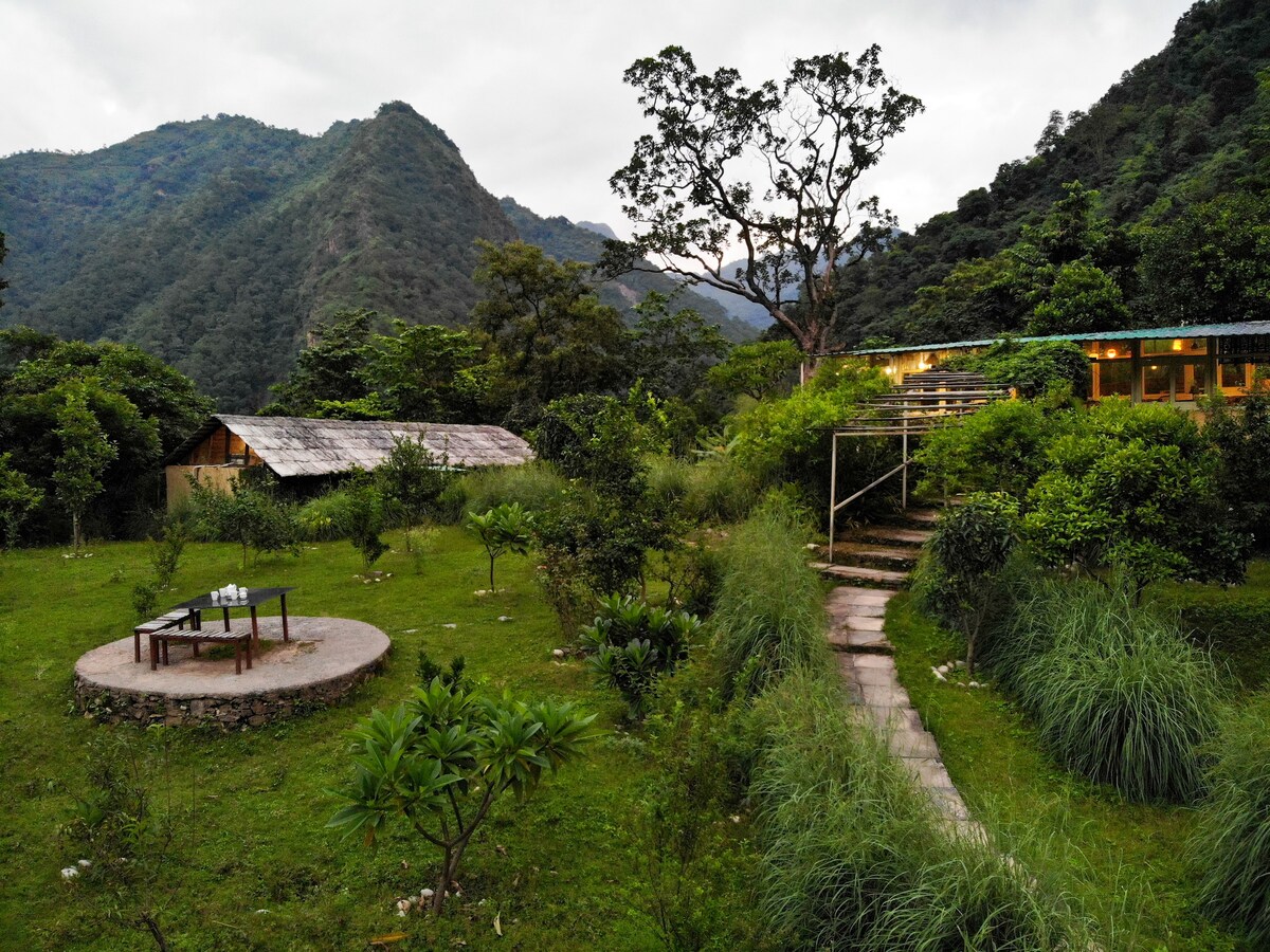A serene landscape is presented, featuring a well-maintained garden surrounded by hills. A circular stone table sits in the foreground, framed by lush greenery. A path leads towards a wooden structure, nestled amid trees, hinting at a peaceful retreat.