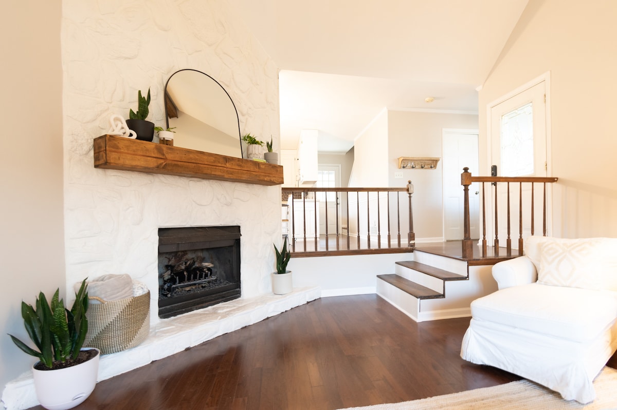 A warm, inviting living area features a white stone fireplace as a focal point, complemented by a wooden shelf with decorative plants. Soft lighting fills the space, and a cozy white chair is situated nearby, adding to the relaxed atmosphere.