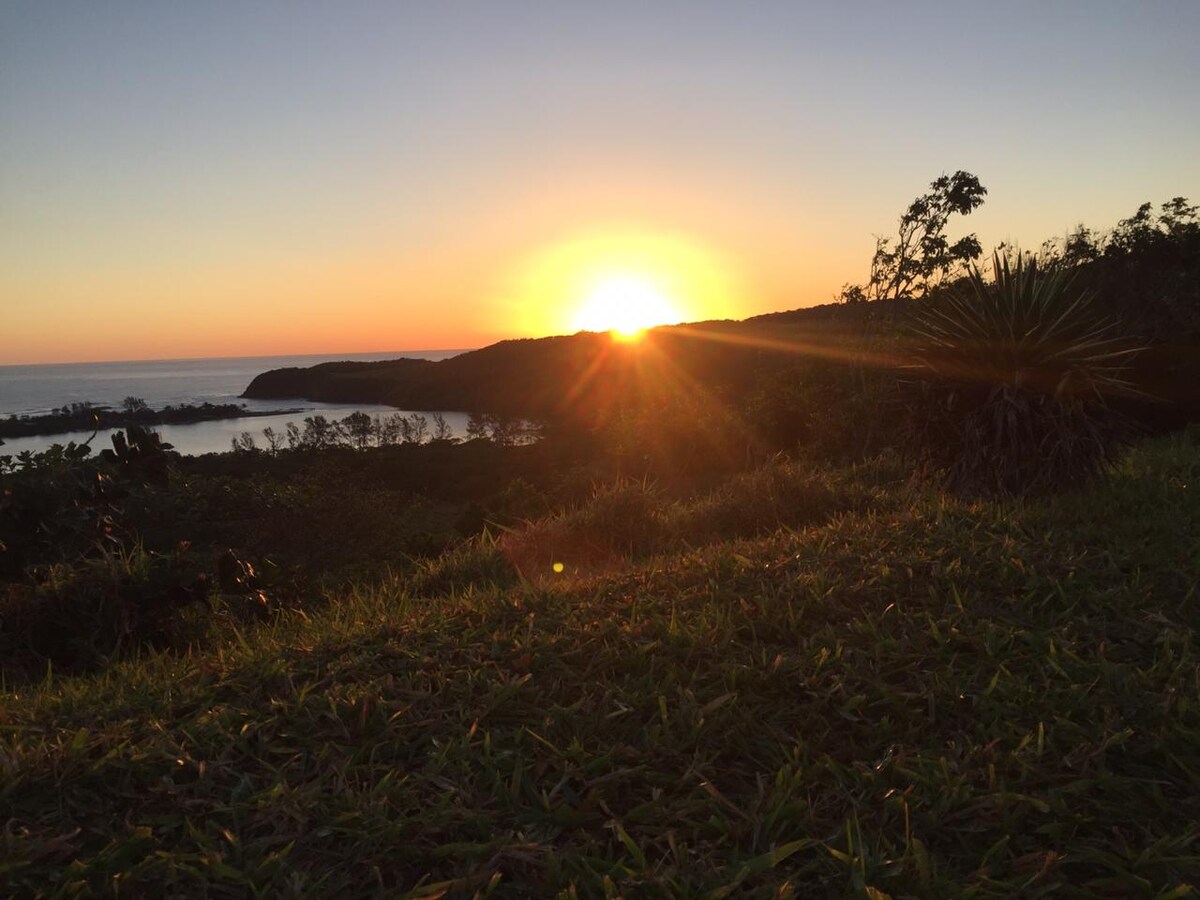 A breathtaking sunset is captured over the horizon, casting warm tones across the sky. The landscape below features lush greenery extending towards the coastline, with water glistening in the fading light.